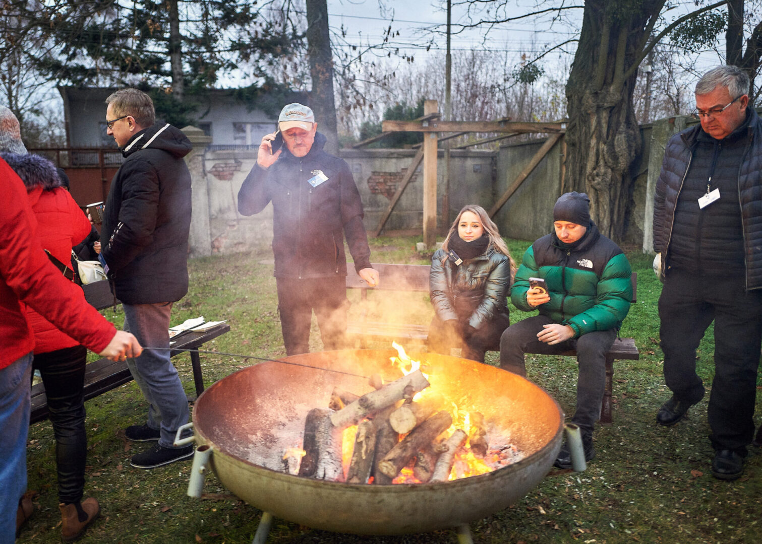 Uczestnicy wycieczki siedzą przy ognisku i pieką kiełbaski. 