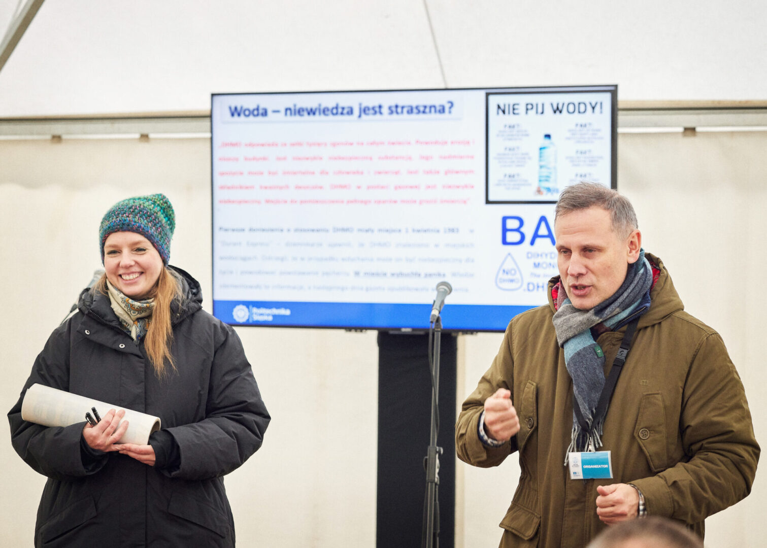 Dyrektor instytucji wraz z pracownicą witają uczestników wycieczki na tle prezentacji pt. "Woda - niewiedza jest straszna?".