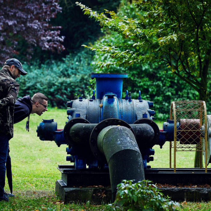 DwĂłch mÄĹźczyzn spoglÄda z zainteresowaniem na zabytkowÄ pompÄ. W tle drzewa i zieleĹ.